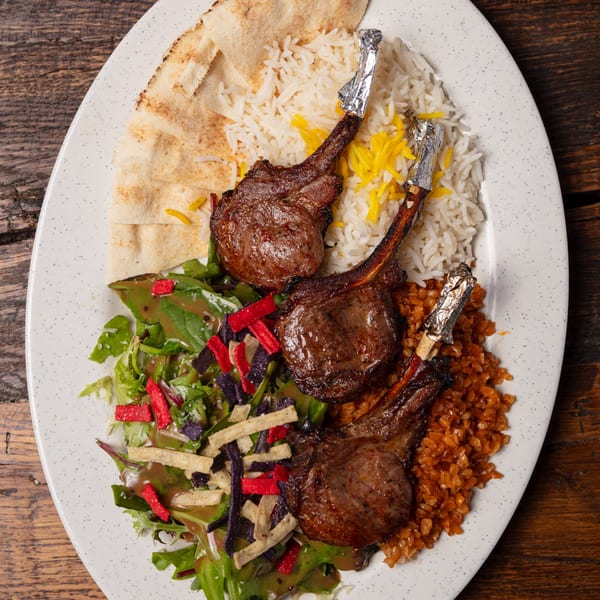 Three lamb chops with rice, salad, and pita bread on a white oval plate.
