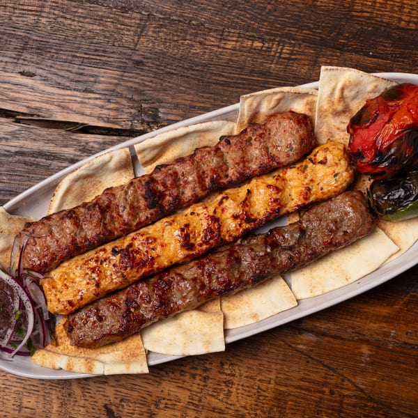 Three skewers of grilled Mediterranean kebabs, pita bread, roasted vegetables, and sliced onions on a white platter.